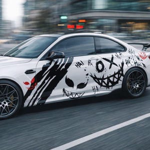May include: A white two-door coupe car featuring black graphic decals on the side panel. The design includes stylized sharp-toothed faces, claw marks, and the repeated text "HA" in a black ink style, with a small red graphic accent near the front.