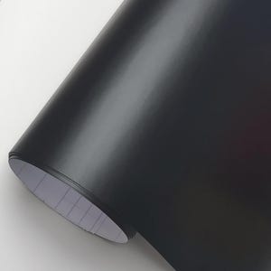 May include: A roll of matte black vinyl material. The material is tightly rolled, revealing a glimpse of the white backing paper. The surface reflects light, showing subtle variations in tone. Ideal for crafting and design projects.
