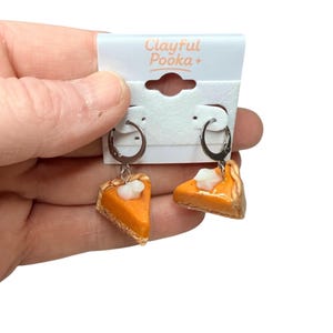 Può includere: Orecchini pendenti con ciondoli a forma di fette di torta di zucca in miniatura. Gli orecchini hanno cerchi argentati e fette di torta arancioni sormontate da panna bianca. Il testo "Clayful Pooka +" è visibile.