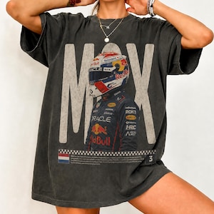May include: A charcoal gray short-sleeve t-shirt featuring a large graphic of a race car driver wearing a helmet and racing suit. The text MAX is printed in large white letters behind the driver, with a checkered flag border at the bottom.