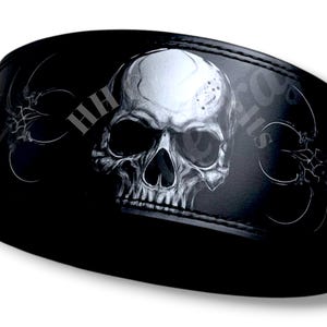 May include: Black weightlifting belt featuring a detailed, grayscale skull design. The belt has a stitched border and decorative patterns on the sides. The text "HH" is visible on the left side.