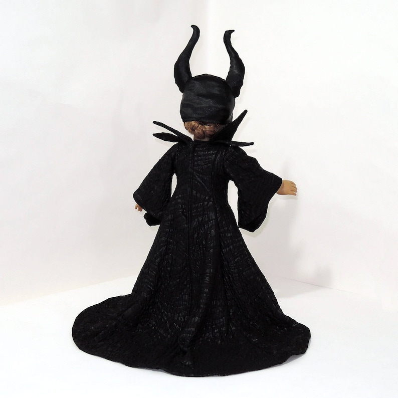 May include: A black doll wearing a black dress with a long train and a black crown with horns. The doll is facing away from the camera.