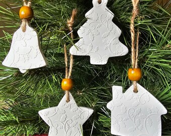 May include: Five white ceramic Christmas ornaments with a floral design hanging on a green pine tree. The ornaments are a bell, a star, a house, and two Christmas trees. Each ornament has a brown twine string with a small yellow bead.