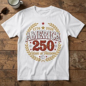 Pode incluir: Uma t-shirt branca de manga curta com um design gráfico contendo o texto AMERICA 250 YEARS OF FREEDOM, 1776 e 2026. O design inclui uma coroa de louros dourada, estrelas vermelhas e brancas e uma estrela dourada central circular.