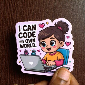Könnte beinhalten: Ein Aufkleber mit einem Cartoon-Mädchen, das auf einem Laptop codiert. Der Aufkleber hat den Text "I CAN CODE my OWN WORLD". Das Mädchen trägt ein orangefarbenes Hemd und rosa Ärmel. Zwei Roboterfiguren befinden sich auf dem Laptop. Rosa Herzen und gelbe Diamanten verzieren den Aufkleber.