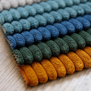Op de afbeelding: Een rechthoekig stoffen staal met een gestructureerd, corduroy-achtig oppervlak met verhoogde, afgeronde horizontale ribbels. Het materiaal vertoont een kleurverloop, variërend van lichtbeige tot blauwgroen, diepblauw, bosgroen en mosterdgeel.