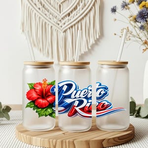 May include: Three frosted glass tumblers with bamboo lids and straws. One features a red hibiscus flower, another displays "Puerto Rico" in blue and red, and the third is plain. The tumblers sit on a wooden tray.
