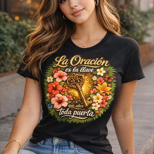 Op de afbeelding: Zwart T-shirt met de Spaanse tekst "La Oración es la llave que abre toda puerta" (Gebed is de sleutel die elke deur opent). Het shirt heeft een sleutelontwerp omringd door bloemen en vlinders.