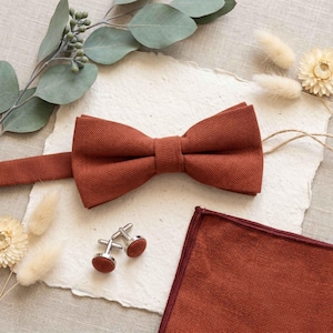 Può includere: Un papillon testurizzato color ruggine, una pochette abbinata con bordo scuro e due gemelli rotondi color argento con inserti in tessuto color ruggine. Gli articoli sono disposti su un foglio di carta testurizzata bianco sporco.