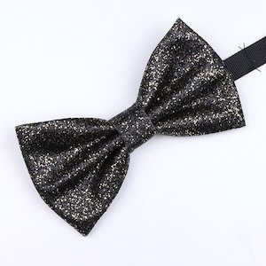 Puede incluir: Una pajarita negra con purpurina y forma de mariposa clásica. La pajarita está hecha de un material brillante y texturizado con motas doradas. La cinta ajustable para el cuello es negra.