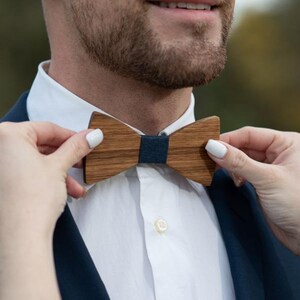 Könnte beinhalten: Eine Holzfliege mit einem dunkelblauen Mittelknoten wird angepasst. Die Fliege ist aus hellbraunem Holz gefertigt. Die Person trägt ein weißes Hemd und einen dunkelblauen Blazer. Die Fliege ist ein stilvolles Accessoire.