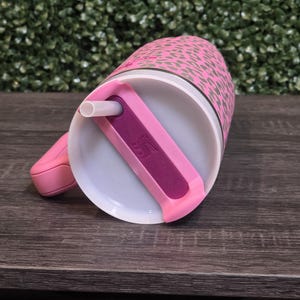 K&ouml;nnte beinhalten: Rosafarbener Becher mit Leopardenmuster. Der Becher hat einen wei&szlig;en Innenraum, einen rosa Griff und einen rosa Deckel mit einem lila Schieber und einem wei&szlig;en Strohhalm. Er steht auf einer dunklen Holzoberfl&auml;che.