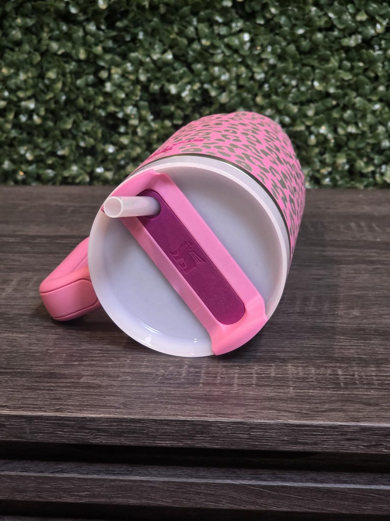 K&ouml;nnte beinhalten: Rosafarbener Becher mit Leopardenmuster. Der Becher hat einen wei&szlig;en Innenraum, einen rosa Griff und einen rosa Deckel mit einem lila Schieber und einem wei&szlig;en Strohhalm. Er steht auf einer dunklen Holzoberfl&auml;che.