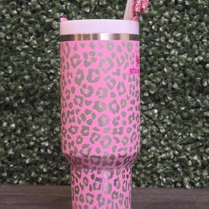 K&ouml;nnte beinhalten: Rosa-grau bedruckter Becher mit Leopardenmuster, wei&szlig;em Deckel und rosa Glitzer-Schleifen-Strohhalm-Aufsatz. Der Edelstahlbecher hat den Aufdruck "STAN" auf der Seite.