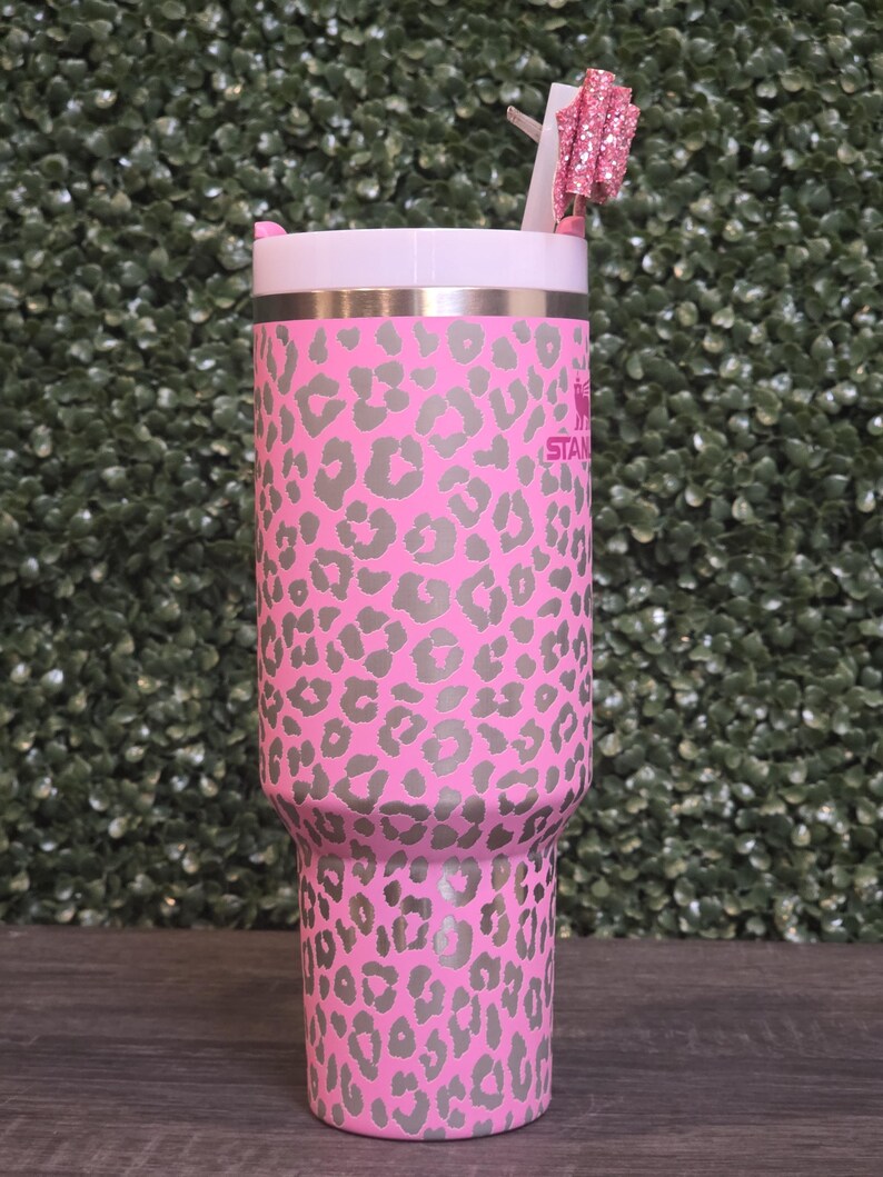 K&ouml;nnte beinhalten: Rosa-grau bedruckter Becher mit Leopardenmuster, wei&szlig;em Deckel und rosa Glitzer-Schleifen-Strohhalm-Aufsatz. Der Edelstahlbecher hat den Aufdruck "STAN" auf der Seite.
