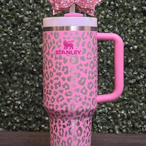 K&ouml;nnte beinhalten: Rosa Stanley-Becher mit grauem Leopardenmuster. Er hat einen rosa Griff und einen wei&szlig;en Deckel. Eine rosa Glitzer-Schleife ist oben befestigt. Das Stanley-Logo ist auf der Vorderseite sichtbar.