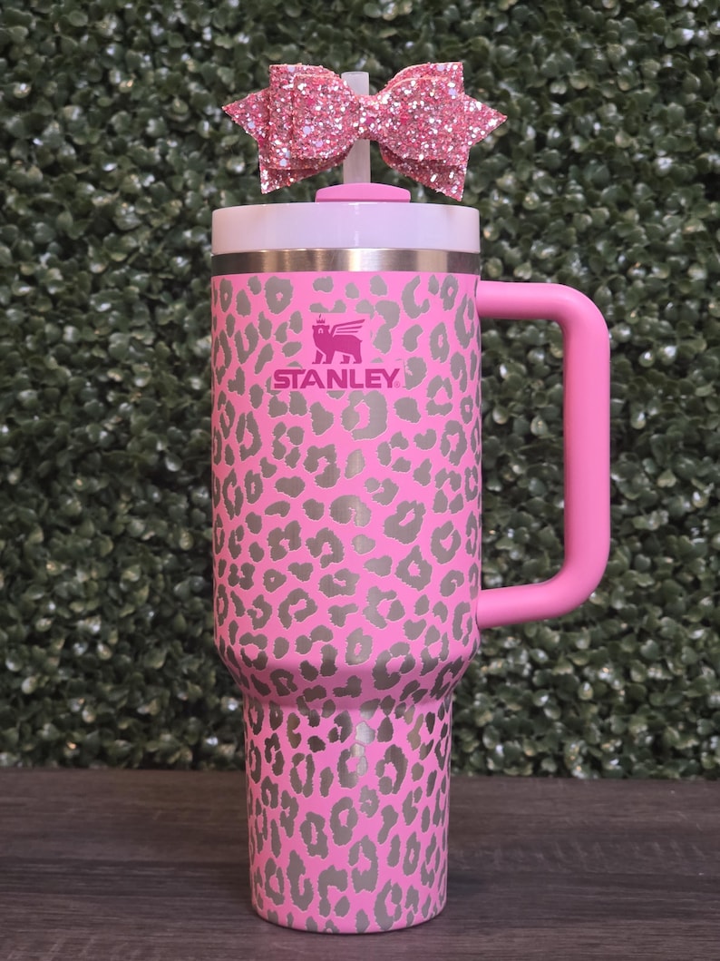K&ouml;nnte beinhalten: Rosa Stanley-Becher mit grauem Leopardenmuster. Er hat einen rosa Griff und einen wei&szlig;en Deckel. Eine rosa Glitzer-Schleife ist oben befestigt. Das Stanley-Logo ist auf der Vorderseite sichtbar.