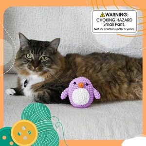 May include: A fluffy brown and white cat rests on a light-colored couch next to a small, handmade purple and white crochet penguin toy. A warning label in the image reads: "CHOKING HAZARD Small Parts. Not for children under 5 years."