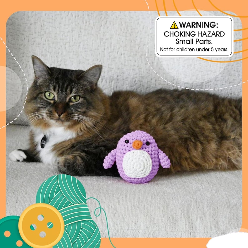 May include: A fluffy brown and white cat rests on a light-colored couch next to a small, handmade purple and white crochet penguin toy. A warning label in the image reads: "CHOKING HAZARD Small Parts. Not for children under 5 years."