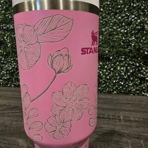 Puede incluir: Un vaso t&eacute;rmico rosa con tapa blanca y pajita. El vaso presenta un dise&ntilde;o floral grabado y la marca "STANLEY" en rosa. El vaso est&aacute; dise&ntilde;ado para mantener las bebidas fr&iacute;as o calientes.