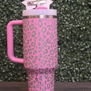 K&ouml;nnte beinhalten: Ein rosa Becher mit grauem Leopardenmuster. Er hat einen rosa Griff und einen wei&szlig;en Deckel. Eine rosa Schleife mit einem silbernen Clip ist am Deckel befestigt. Der Becher ist etwa 23 cm hoch.