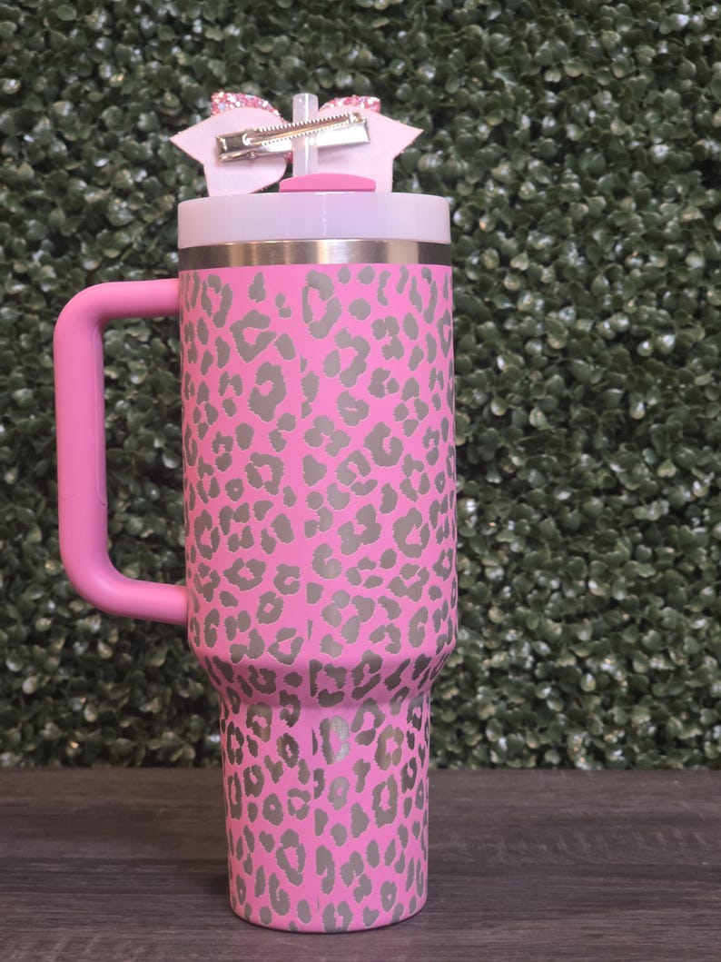 K&ouml;nnte beinhalten: Ein rosa Becher mit grauem Leopardenmuster. Er hat einen rosa Griff und einen wei&szlig;en Deckel. Eine rosa Schleife mit einem silbernen Clip ist am Deckel befestigt. Der Becher ist etwa 23 cm hoch.