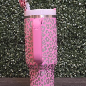 K&ouml;nnte beinhalten: Rosafarbener Becher mit grauem Leopardenmuster. Der Becher hat einen rosa Griff, einen wei&szlig;en Deckel und einen silbernen Rand. Ein rosa Glitzerstift und ein wei&szlig;er Strohhalm sind in den Deckel eingesetzt. Der Becher steht auf einer Holzoberfl&auml;che.