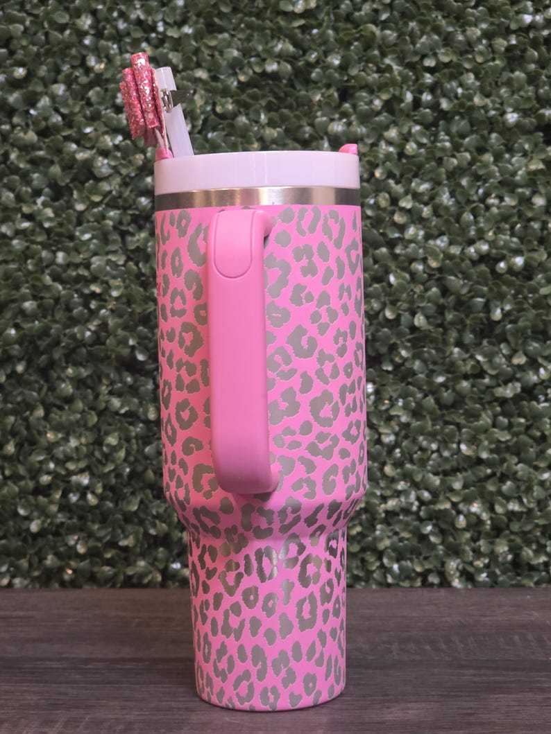 K&ouml;nnte beinhalten: Rosafarbener Becher mit grauem Leopardenmuster. Der Becher hat einen rosa Griff, einen wei&szlig;en Deckel und einen silbernen Rand. Ein rosa Glitzerstift und ein wei&szlig;er Strohhalm sind in den Deckel eingesetzt. Der Becher steht auf einer Holzoberfl&auml;che.