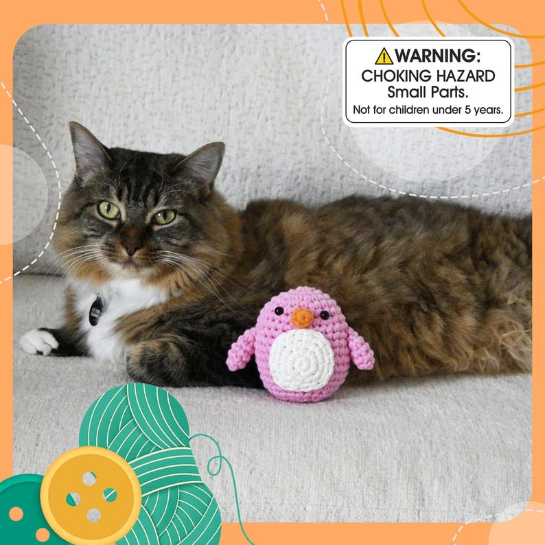May include: A fluffy brown and white cat rests near a pink and white crocheted penguin toy. The toy has a white belly, orange beak, and black eyes. A warning label about choking hazards is visible.