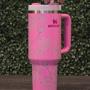Puede incluir: Un vaso Stanley rosa con un dise&ntilde;o floral y un asa rosa. El vaso tiene una tapa blanca y un lazo rosa en la parte superior. El logotipo de Stanley es visible en el lateral.