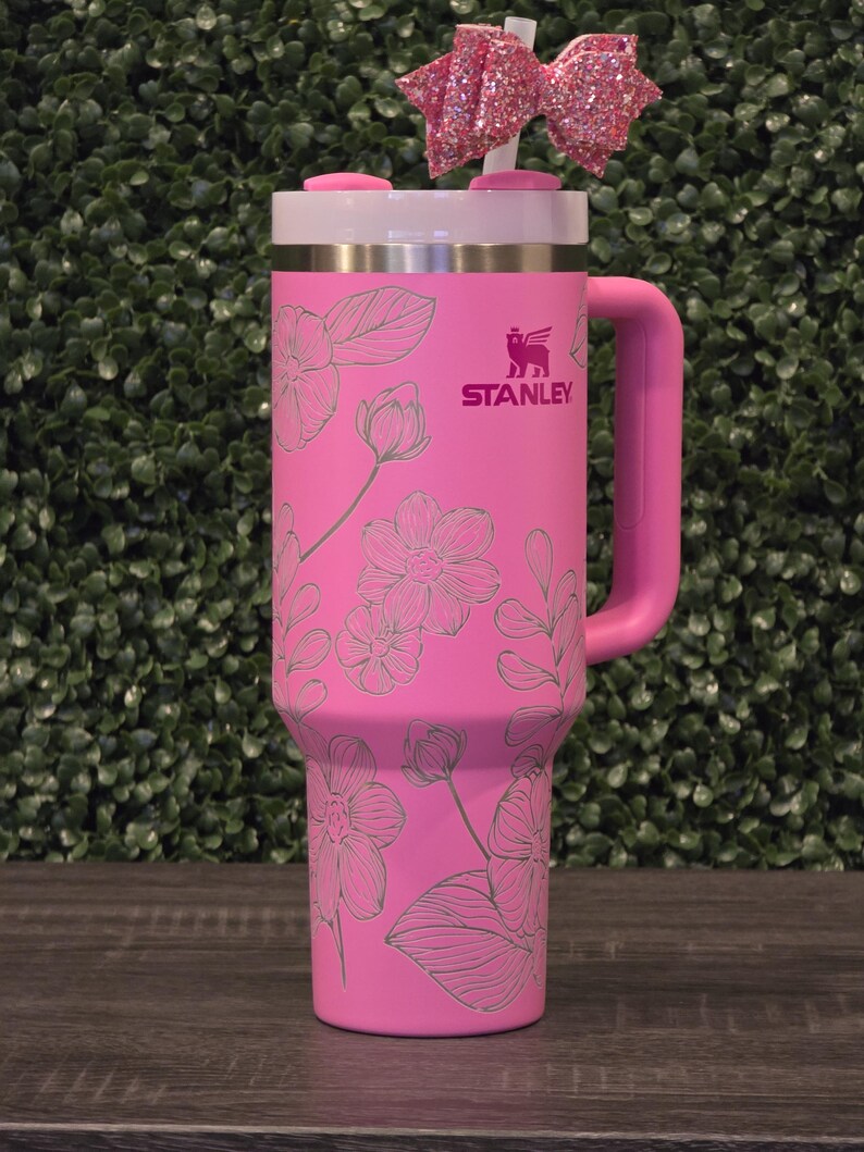 Puede incluir: Un vaso Stanley rosa con un dise&ntilde;o floral y un asa rosa. El vaso tiene una tapa blanca y un lazo rosa en la parte superior. El logotipo de Stanley es visible en el lateral.