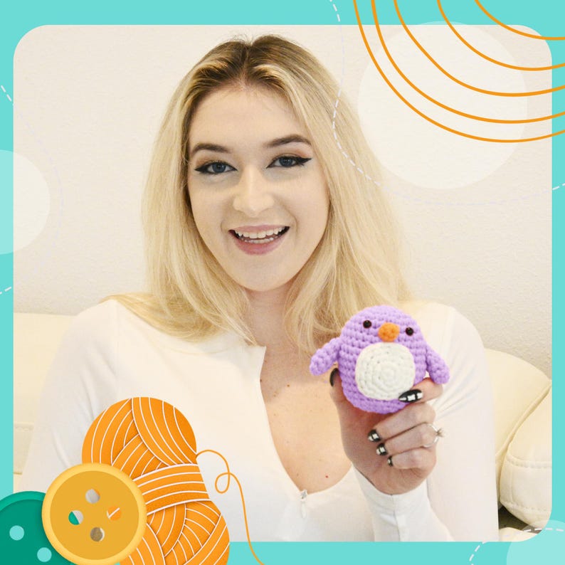 May include: A small, handmade crochet penguin in shades of purple, white, and orange. The penguin has a round body, small wings, and a white belly. The penguin is held in a person's hand.