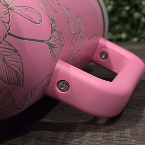 Puede incluir: Un vaso t&eacute;rmico rosa con un asa a juego. El vaso presenta un dise&ntilde;o floral en un tono rosa m&aacute;s claro. El asa est&aacute; sujeta con tornillos plateados. El vaso est&aacute; dise&ntilde;ado para mantener las bebidas calientes o fr&iacute;as.