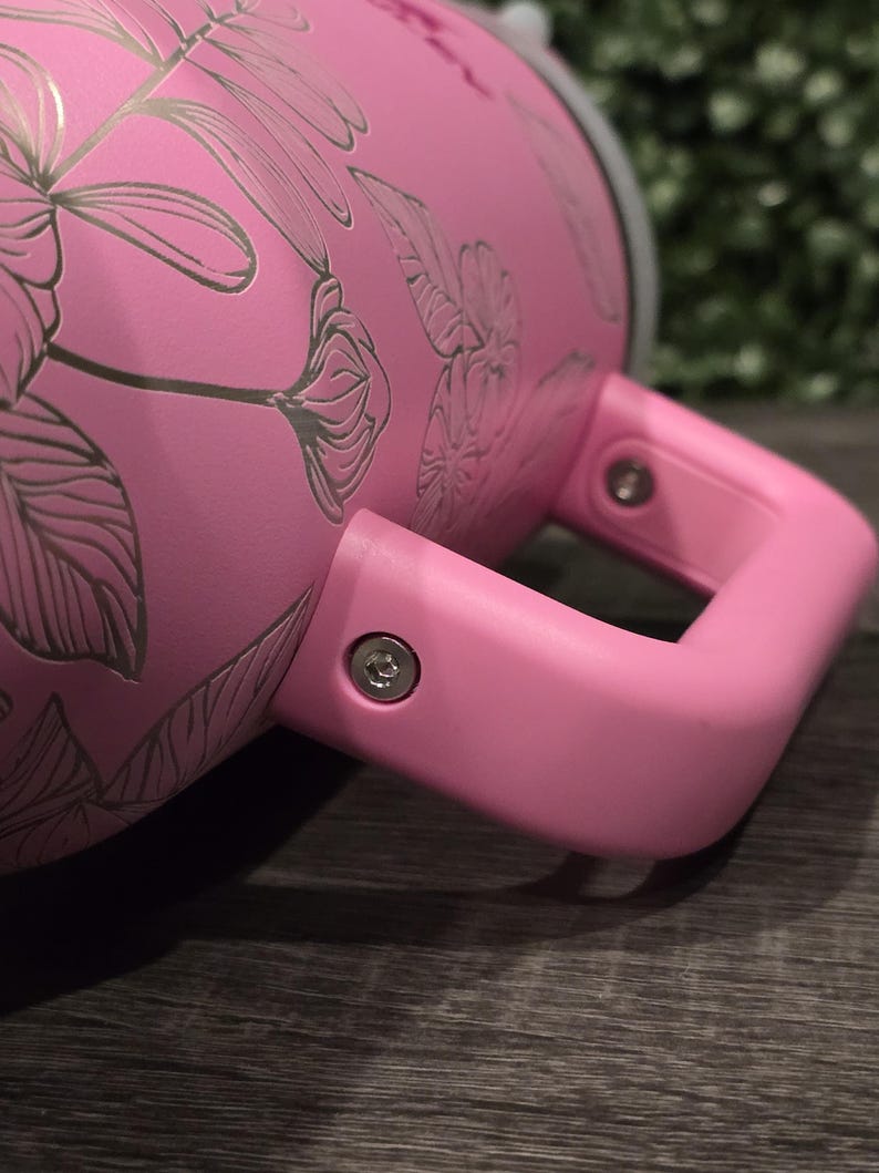 Puede incluir: Un vaso t&eacute;rmico rosa con un asa a juego. El vaso presenta un dise&ntilde;o floral en un tono rosa m&aacute;s claro. El asa est&aacute; sujeta con tornillos plateados. El vaso est&aacute; dise&ntilde;ado para mantener las bebidas calientes o fr&iacute;as.