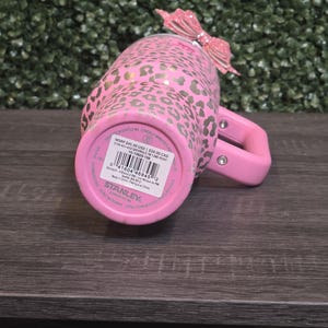K&ouml;nnte beinhalten: Rosa Stanley-Becher mit goldenem Leopardenmuster und rosa Griff. Oben ist eine rosa Glitzer-Schleife befestigt. Auf der Unterseite des Bechers sind der Markenname und Produktinformationen zu sehen.