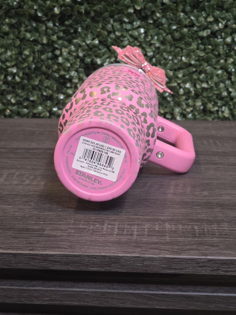 K&ouml;nnte beinhalten: Rosa Stanley-Becher mit goldenem Leopardenmuster und rosa Griff. Oben ist eine rosa Glitzer-Schleife befestigt. Auf der Unterseite des Bechers sind der Markenname und Produktinformationen zu sehen.