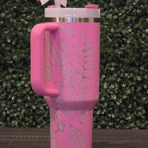 Puede incluir: Un vaso t&eacute;rmico rosa con asa, adornado con un dise&ntilde;o floral. El vaso tiene una tapa blanca, un asa rosa y una pajita. Un lazo decorativo est&aacute; sujeto a la pajita. Dise&ntilde;ado para mantener las bebidas fr&iacute;as o calientes.