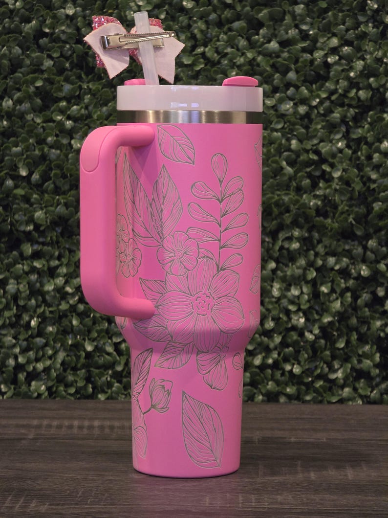 Puede incluir: Un vaso t&eacute;rmico rosa con asa, adornado con un dise&ntilde;o floral. El vaso tiene una tapa blanca, un asa rosa y una pajita. Un lazo decorativo est&aacute; sujeto a la pajita. Dise&ntilde;ado para mantener las bebidas fr&iacute;as o calientes.
