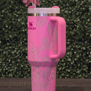 Puede incluir: Un vaso Stanley rosa con un dise&ntilde;o floral y un asa. El vaso tiene una tapa blanca y un lazo rosa en la pajita. El logotipo de Stanley es visible en la parte delantera.