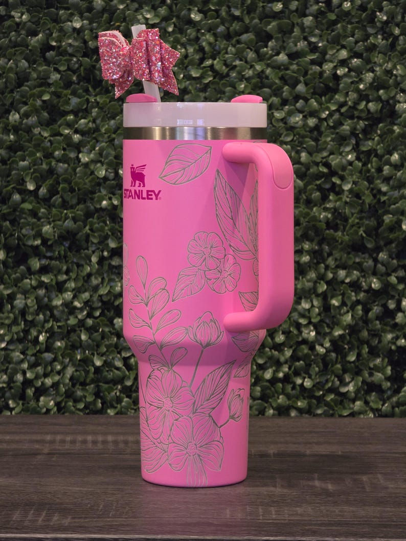 Puede incluir: Un vaso Stanley rosa con un dise&ntilde;o floral y un asa. El vaso tiene una tapa blanca y un lazo rosa en la pajita. El logotipo de Stanley es visible en la parte delantera.