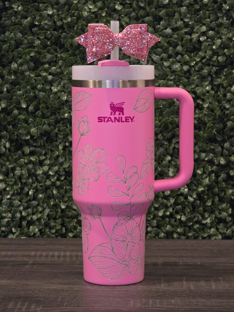 Puede incluir: Un vaso Stanley rosa con un dise&ntilde;o floral y un asa a juego. El vaso tiene una tapa blanca y un lazo rosa brillante en la parte superior. El logotipo de Stanley es visible en la parte delantera.