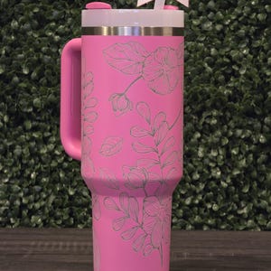 Puede incluir: Un vaso t&eacute;rmico rosa con asa, con un dise&ntilde;o floral. El vaso tiene una tapa blanca, una banda plateada y un asa rosa. Una pajita blanca con un lazo rosa est&aacute; insertada en la tapa. El fondo es un seto verde.