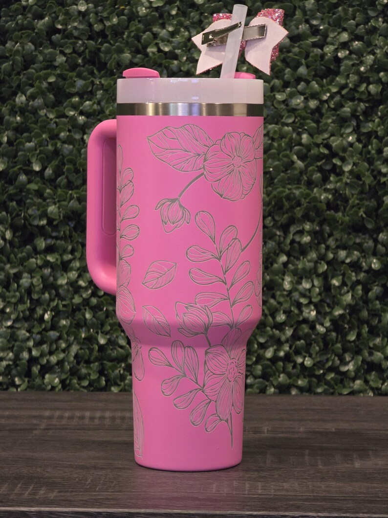 Puede incluir: Un vaso t&eacute;rmico rosa con asa, con un dise&ntilde;o floral. El vaso tiene una tapa blanca, una banda plateada y un asa rosa. Una pajita blanca con un lazo rosa est&aacute; insertada en la tapa. El fondo es un seto verde.
