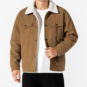 Op de afbeelding: Een bruine corduroy jas met een witte, donzige kraag en voering. De jas heeft twee borstzakken en zwarte knopen. De mouwen hebben manchetten met knopen. De jas wordt over een wit t-shirt gedragen.