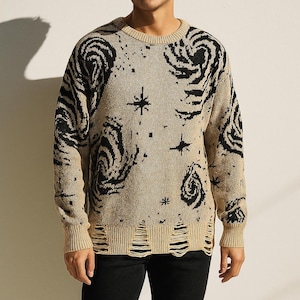 Puede incluir: Suéter beige con patrones de espirales y estrellas negras. El suéter tiene cuello redondo, mangas largas y detalles desgastados en el dobladillo. El diseño le da al suéter un aspecto artístico único.