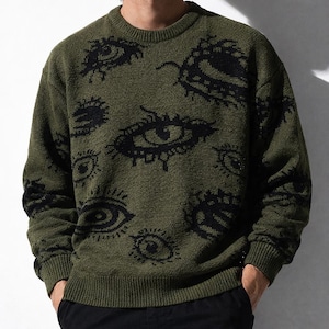 Pode incluir: Camisola verde-oliva com gola redonda e um padrão repetido de olhos pretos. A camisola tem gola, punhos e bainha canelados. O design apresenta olhos de vários tamanhos e estilos, criando um visual artístico único. A camisola é feita de um material de malha macia.