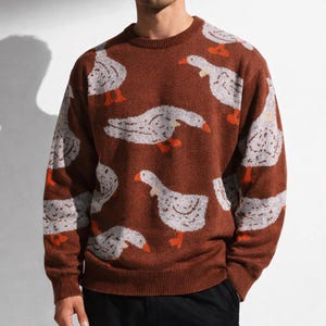 May include: A brown knit sweater featuring a repeating pattern of white geese with orange feet and beaks. The sweater has a crew neck and long sleeves. The geese are in various poses, creating a playful and whimsical design. This cozy sweater is perfect for casual wear.