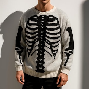 Op de afbeelding: Een lichtgrijze trui met een zwart skeletontwerp op de voorkant en mouwen. De trui heeft een ronde hals en geribde manchetten en zoom. Het skeletontwerp omvat de ribbenkast, wervelkolom en armbotten.