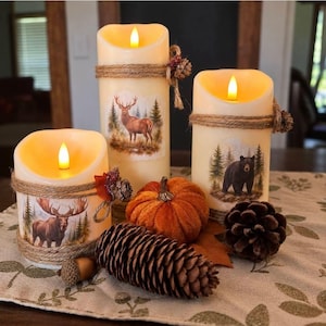 May include: Three cream-colored LED pillar candles featuring wildlife illustrations of a moose, a deer, and a black bear. Each candle is wrapped with twine and accented with small pinecones. The set is displayed with a fabric pumpkin and pinecones.