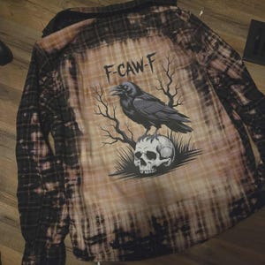 May include: A long-sleeved, button-down flannel shirt with a bleached, distressed design. The back features a graphic of a raven perched on a skull, with the text "F-CAW-F" above. The shirt has a plaid pattern in black, brown, and tan.